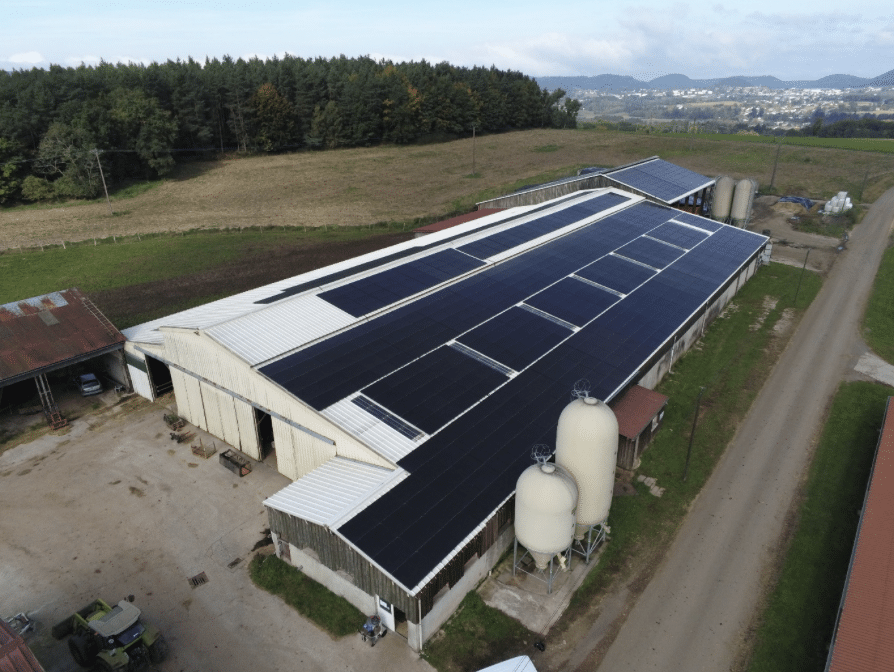 Une grande grange dont le toit est recouvert de panneaux solaires est entourée de champs, de silos et de bâtiments proches, avec une route le long de la route et une forêt et des collines à l'arrière-plan.