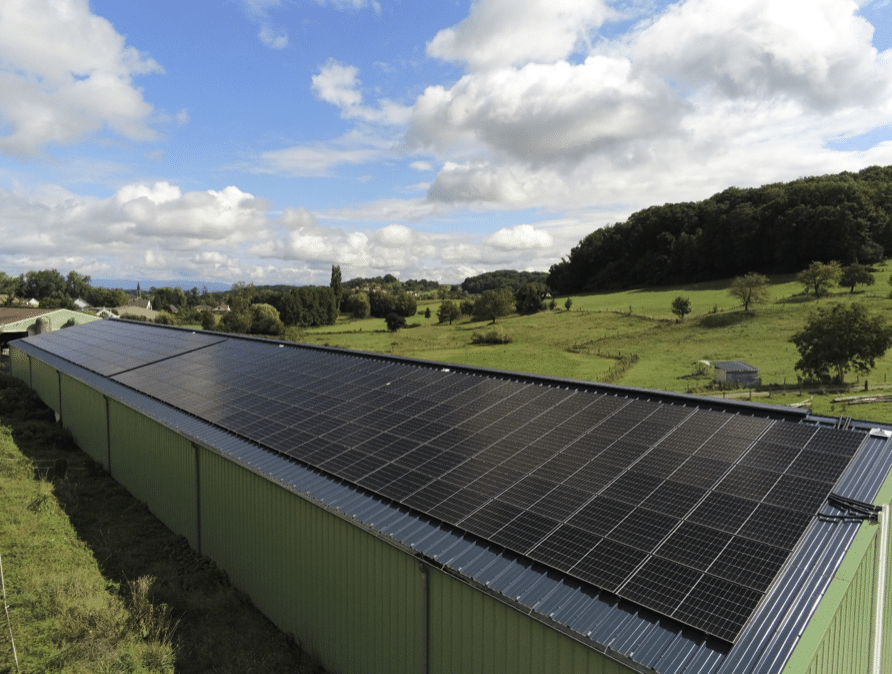 Des panneaux solaires recouvrent un grand bâtiment vert situé dans une zone rurale avec des champs herbeux, des arbres épars et des collines lointaines, sous un ciel partiellement nuageux.