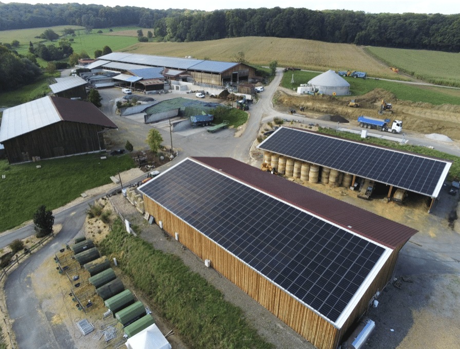 Vue aérienne d'une ferme rurale comprenant plusieurs bâtiments, dont la plupart sont équipés de grands panneaux solaires installés sur leurs toits. La ferme est entourée de champs, d'arbres et de divers équipements agricoles.