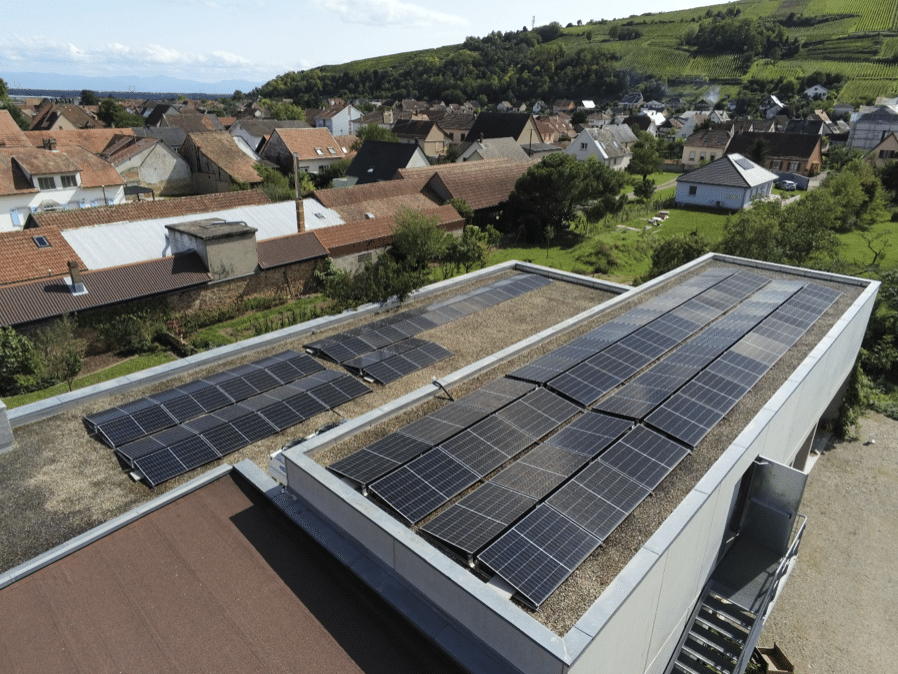 Vue aérienne de toits équipés de panneaux solaires installés sur des bâtiments plats dans un village, avec des maisons, de la verdure et des collines en arrière-plan, sous un ciel partiellement nuageux.