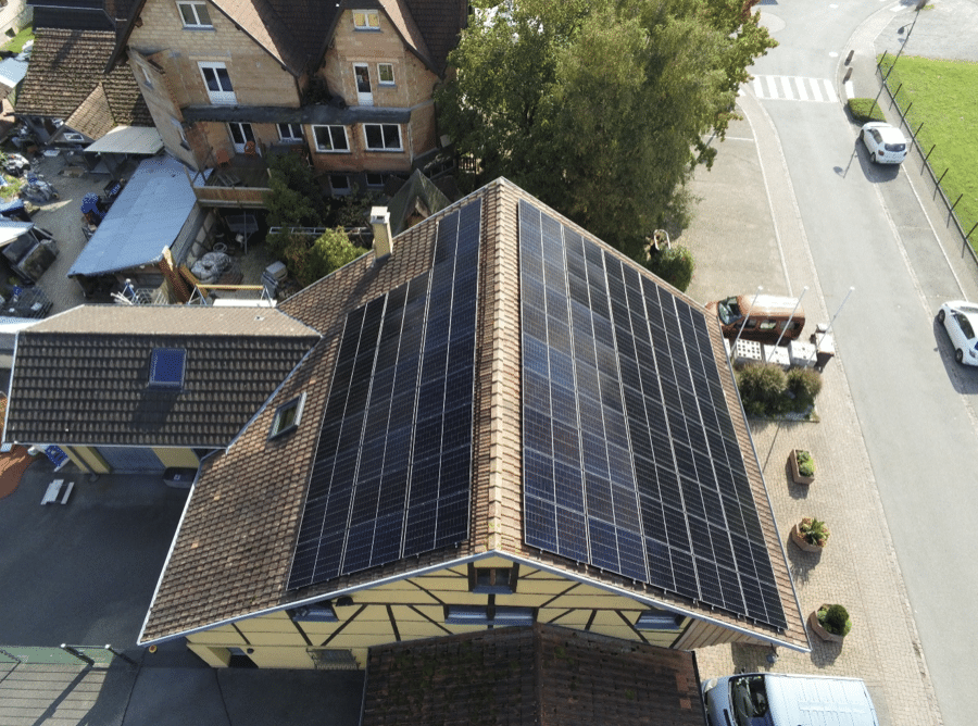 Vue aérienne d'une maison au toit de tuiles brunes couverte de panneaux solaires, entourée d'autres bâtiments, de verdure et de voitures garées le long d'une route voisine.