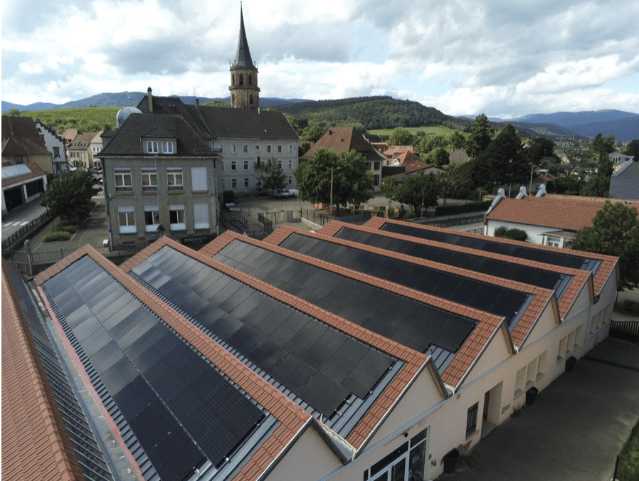 Une rangée de bâtiments avec de grands panneaux solaires sur leurs toits, situés dans une petite ville européenne avec un clocher d'église, des maisons et des collines vertes visibles en arrière-plan sous un ciel partiellement nuageux.